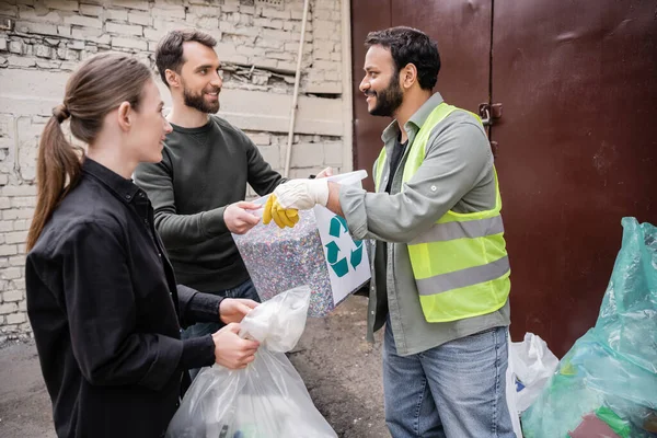 Voluntario sonriente dando papelera con signo de reciclaje al trabajador indio en chaleco de seguridad y guantes al aire libre en la estación de eliminación de residuos, clasificación de basura y concepto de reciclaje - foto de stock