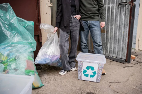 Cropped view of volunteers standing near trash bin and bags near door of waste disposal station outdoors, garbage sorting and recycling concept — Stock Photo