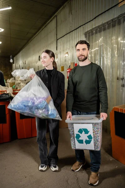 Positive volunteers holding trash bin and bag while standing together in blurred waste disposal station at background, garbage sorting and recycling concept — Stock Photo