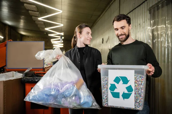 Smiling volunteer holding trash bag near man with bin and recycle sign in blurred waste disposal station at background, garbage sorting and recycling concept — Stock Photo