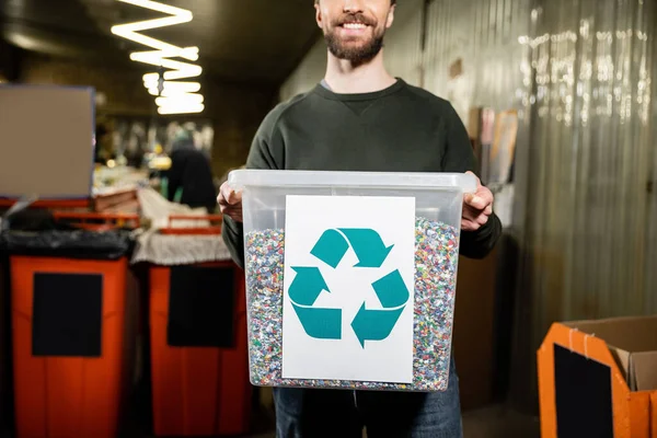 Vista recortada del voluntario sonriente y barbudo sosteniendo la papelera con el cartel de reciclaje en la estación de eliminación de residuos borrosa en el fondo, clasificación de la basura y el concepto de reciclaje - foto de stock