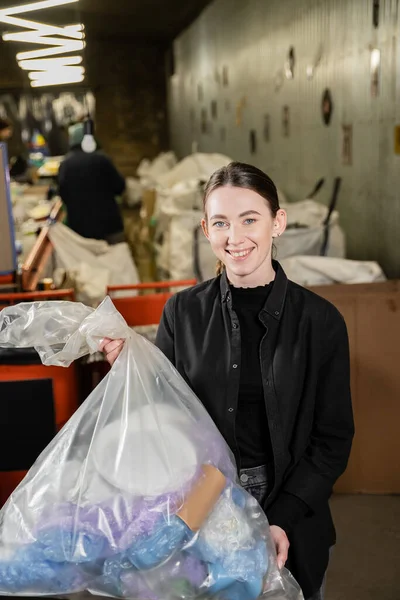 Positive female volunteer looking at camera while holding plastic bag with trash in blurred waste disposal station at background, garbage sorting and recycling concept — Stock Photo