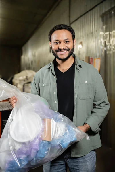 Lächelnder indischer Freiwilliger blickt in die Kamera, während er im Hintergrund eine Plastiktüte mit Müll in einer unscharfen Müllentsorgungsstation hält, Mülltrennung und Recyclingkonzept — Stockfoto