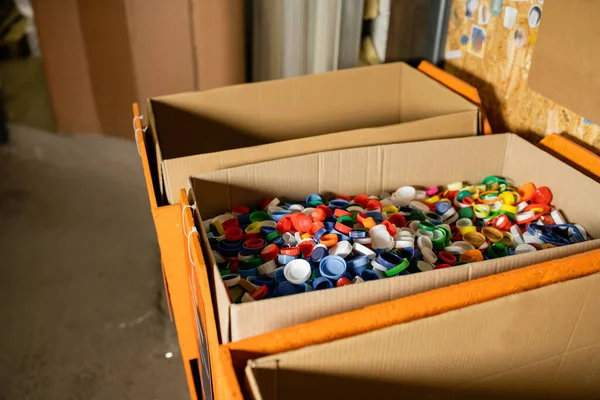 Cardboard boxes with colorful plastic caps for recycle in blurred waste disposal station at background, garbage sorting and recycling concept — Stock Photo