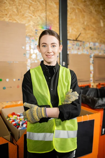 Smiling worker in high visibility vest and gloves looking at camera and crossing arms while standing near blurred plastic caps in box waste disposal station, garbage sorting and recycling concept — Stock Photo