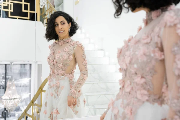 Брюнетка Среднего Востока женщина с волнистыми волосами, стоящие в великолепном и цветочном свадебном платье, глядя на зеркало в роскошном свадебном салоне, счастливая невеста, очаровательный и элегантный, отражение, покупки — стоковое фото