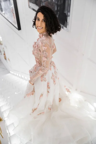 Morena do Oriente Médio mulher com cabelo ondulado de pé em vestido de noiva lindo e floral, enquanto sorrindo no luxuoso salão de noiva e olhando para a câmera, noiva feliz, encantador e elegante, compras — Fotografia de Stock