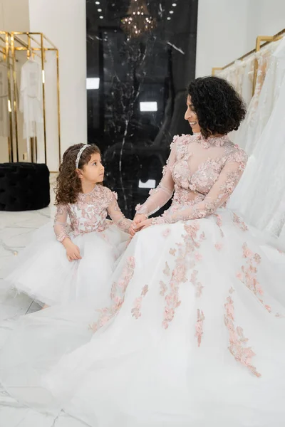 Happy and brunette middle eastern woman with wavy hair looking at cute girl and smiling near white wedding dresses in bridal salon, floral, mother and daughter, happiness, wedding day, shopping — Stock Photo