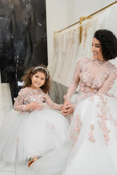 Брюнетка Ближнего Востока женщина с волнистыми волосами глядя на веселую девушку и улыбаясь возле белых свадебных платьев в свадебном салоне, цветочный, мать и дочь, счастье, покупки, день свадьбы — стоковое фото