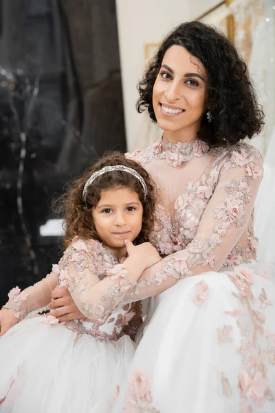 Brunette middle eastern woman with wavy hair hugging cute girl and smiling near white wedding dresses in bridal salon, floral, mother and daughter, happiness, shopping, wedding day — Stock Photo