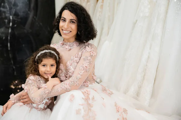 Brunette middle eastern woman with wavy hair embracing cute girl and smiling near white wedding dresses in bridal boutique, floral, mother and daughter, happiness, wedding day — Stock Photo