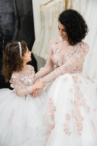 Brunette femme du Moyen-Orient avec les cheveux ondulés regardant fille et souriant près des robes de mariée blanches dans le salon de mariée, floral, mère et fille, bonheur, jour du mariage, shopping, liaison — Photo de stock