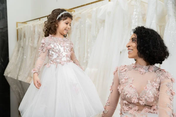 Joyful middle eastern woman with wavy hair looking at cute girl and smiling near white wedding dresses in bridal salon, floral, mother and daughter, happiness, wedding day, shopping — Stock Photo