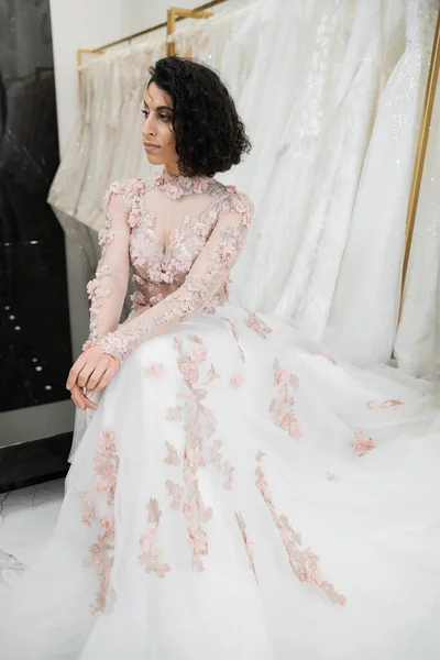 Брюнетка Среднего Востока женщина с волнистыми волосами сидя в роскошном и цветочном свадебном платье рядом размытое и белое платье внутри роскошного свадебного салона, очаровательный и элегантный, невеста-для-быть, покупки — стоковое фото