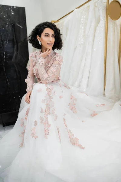 Mulher feliz do Oriente Médio com cabelo ondulado sentado em vestido de noiva lindo e floral perto de vestido borrado e branco dentro do luxuoso salão de noiva, charmoso e elegante, noiva-a-ser — Fotografia de Stock