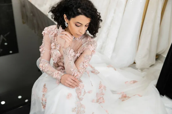 Vue aérienne de la femme brune du Moyen-Orient avec des cheveux ondulés assis dans une robe de mariée magnifique et florale près de robe floue et blanche à l'intérieur du luxueux salon de mariée, shopping, future mariée — Photo de stock