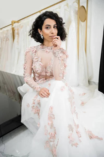 Brunette middle eastern woman with wavy hair sitting in gorgeous and floral wedding dress near blurred and white gown inside of luxurious bridal salon, shopping, bride-to-be, looking at camera — Stock Photo