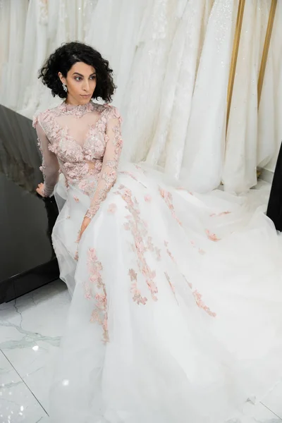 Morena do Oriente Médio mulher com cabelo ondulado sentado em vestido de noiva lindo e floral com trem perto de vestidos borrados e brancos dentro do luxuoso salão de noiva, noiva-a-ser — Fotografia de Stock