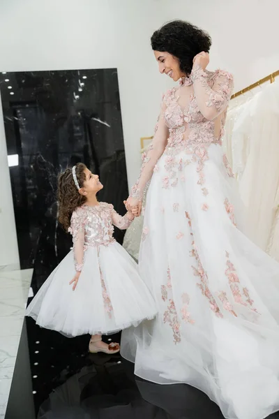Cheerful middle eastern woman with wavy hair in stunning wedding dress holding hands with daughter in cute floral attire while standing in bridal salon, shopping, luxurious, golden accents — Stock Photo