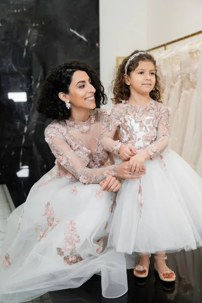 Brunette middle eastern woman with wavy hair in stunning wedding dress holding hand of cute daughter in cute floral attire in bridal salon, shopping, happiness, special moment, togetherness — Stock Photo