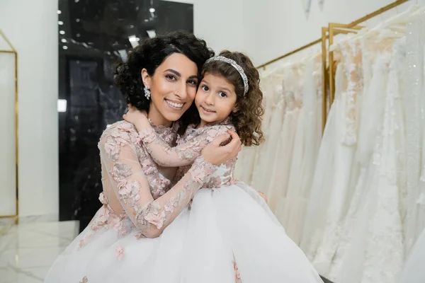 Brunette middle eastern woman with wavy hair in stunning wedding dress hugging cute daughter in cute floral attire in bridal salon, shopping, happiness, special moment, togetherness — Stock Photo
