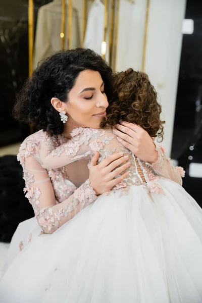 Очаровательный ближневосточная невеста с волнистыми волосами в потрясающем свадебном платье обнимая дочь в милой цветочной одежде в свадебном салоне, покупки, счастье, особый момент, единение, блаженство — стоковое фото