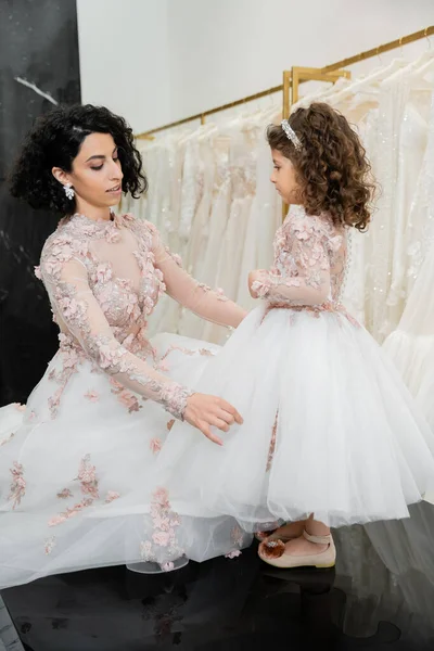 Charming middle eastern bride with brunette wavy hair in wedding dress adjusting tulle skirt of daughter in cute floral attire in bridal salon, shopping, special moment, togetherness — Stock Photo