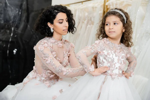 Alluring middle eastern bride with brunette wavy hair in wedding dress adjusting cute floral dress of daughter in bridal salon, shopping, special moment, hands on hips, blurred white gown — Stock Photo