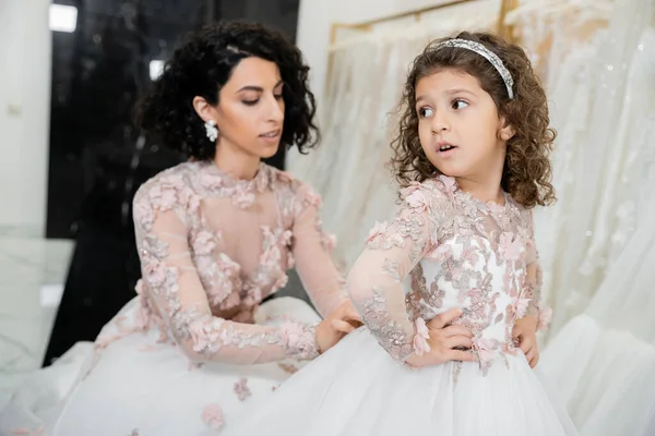 Blurred middle eastern bride with brunette wavy hair in wedding dress adjusting cute floral dress of surprised daughter in bridal salon, shopping, special moment, hands on hips, white gown — Stock Photo