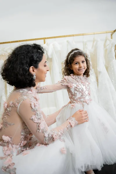Happy middle eastern woman with brunette wavy hair in floral wedding dress adjusting tulle skirt of daughter in cute attire in bridal salon, shopping, special moment, togetherness — Stock Photo