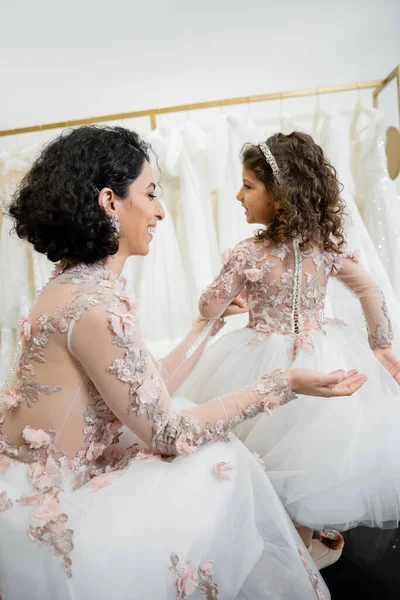 Happy middle eastern woman with brunette wavy hair in floral wedding dress looking at tulle skirt of smiling daughter in cute attire in bridal salon, shopping, special moment, togetherness — Stock Photo