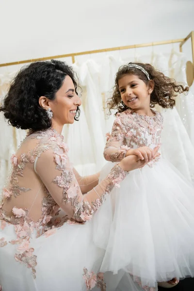 Happy middle eastern bride with brunette wavy hair in floral wedding dress holding hands with smiling daughter in cute attire with tulle skirt in bridal salon, shopping, special moment — Stock Photo