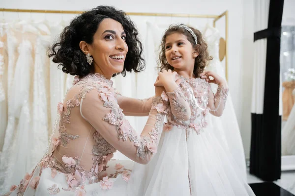 Happy middle eastern woman with brunette hair in floral wedding dress hugging smiling daughter in cute attire with tulle skirt in bridal salon, shopping, special moment, togetherness, looking away — Stock Photo