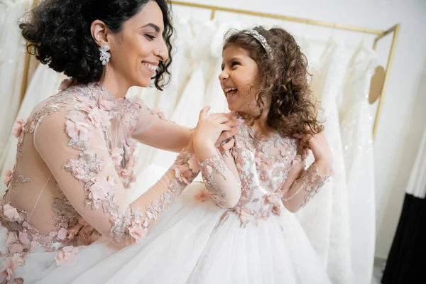 Happy middle eastern woman with brunette hair in floral wedding dress hugging shoulders of smiling daughter in cute attire with tulle skirt in bridal salon, shopping, special moment, togetherness — Stock Photo