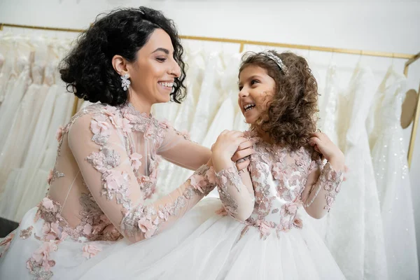 Happy middle eastern woman in floral wedding dress hugging shoulders of smiling girl in cute attire with tulle skirt in bridal salon, shopping, special moment, mother and daughter — Stock Photo
