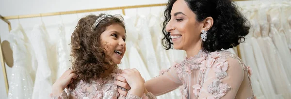 Happy middle eastern woman in floral wedding dress hugging shoulders of cheerful girl in cute attire in bridal salon, shopping, special moment, mother and daughter, bonding, banner — Stock Photo
