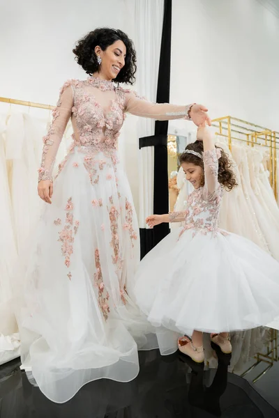 Счастливая женщина с Ближнего Востока в цветочном свадебном платье танцует с улыбающейся девушкой в милой одежде с тюлем юбка в свадебный салон, покупки, особый момент, мать и дочь, счастье — стоковое фото