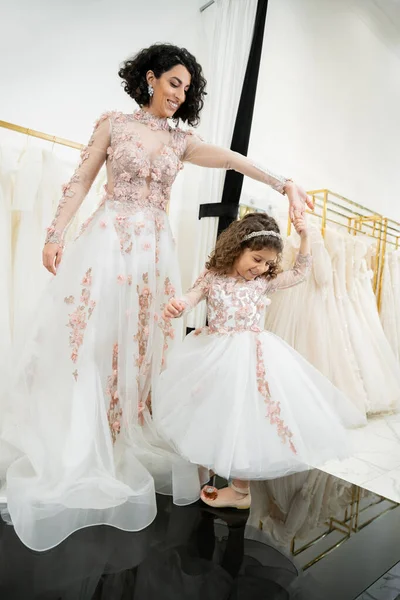 Счастливая женщина с Ближнего Востока в цветочном свадебном платье танцует с улыбающейся девушкой в милой одежде с тюлем юбка в свадебный салон, покупки, особый момент, мать и дочь, счастье — стоковое фото