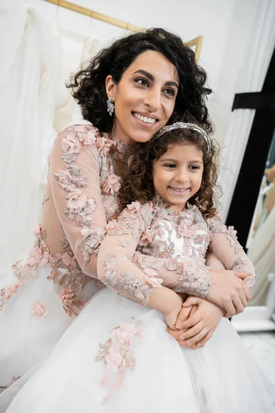 Fröhliche Frau aus dem Nahen Osten im floralen Hochzeitskleid umarmt glückliches Mädchen in netter Kleidung im Brautsalon, Shopping, besonderer Moment, Mutter und Tochter, Glück — Stockfoto