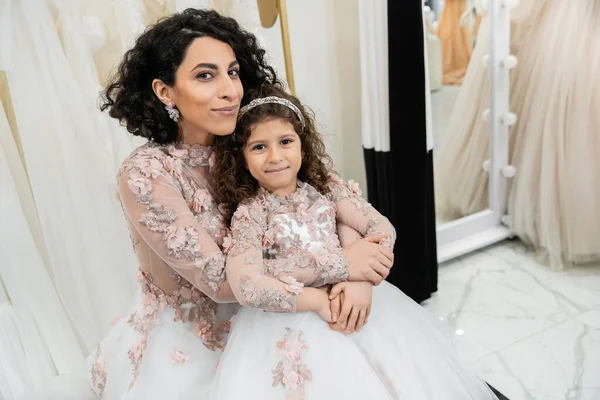 Encantadora mulher do Oriente Médio em vestido de casamento floral abraçando menina feliz em traje bonito no salão de noiva, compras, momento especial, mãe e filha, felicidade, olhando para a câmera — Fotografia de Stock