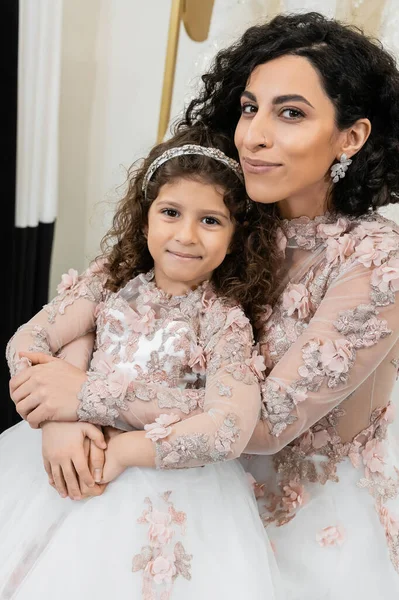 Cheerful middle eastern bride in wedding dress hugging happy girl in cute floral attire in bridal salon, shopping, special moment, mother and daughter, happiness, looking at camera — Stock Photo