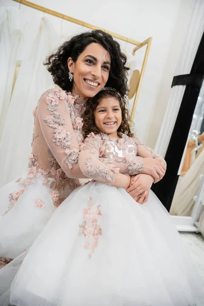 Delightful middle eastern bride in wedding dress hugging happy girl in cute floral attire in bridal salon, shopping, special moment, mother and daughter, happiness, looking away — Stock Photo