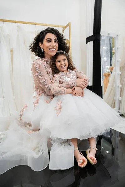 Joyful middle eastern bride in floral wedding dress hugging happy girl in cute attire in bridal salon, shopping, special moment, mother and daughter, happiness, looking away, white gown — Stock Photo