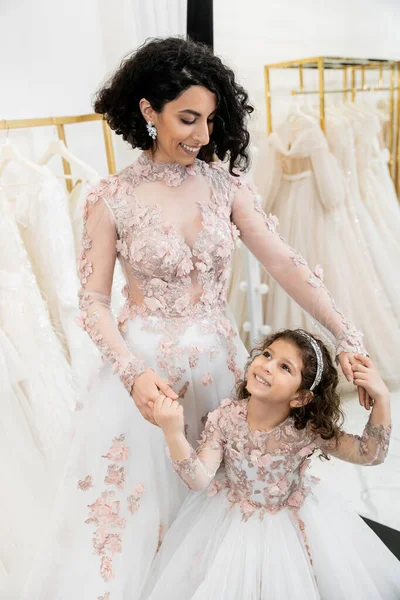 Delightful middle eastern bride in floral wedding dress holding hands with happy girl in cute attire in bridal salon, shopping, special moment, mother and daughter, happiness, togetherness — Stock Photo
