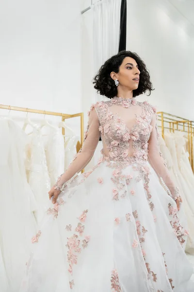 Enchanting and middle eastern woman trying on gorgeous and floral wedding dress inside of luxurious bridal salon, shopping, bride-to-be,  blurred white gown on background, looking away — Stock Photo
