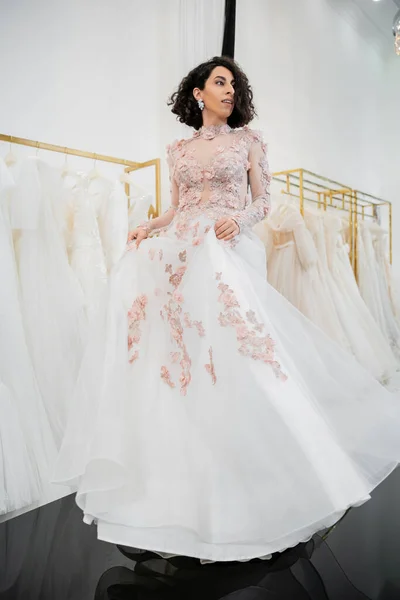 Full length of brunette and middle eastern woman with wavy hair trying on gorgeous and floral wedding dress inside of luxurious bridal salon, shopping, bride-to-be,  blurred white gown — Stock Photo