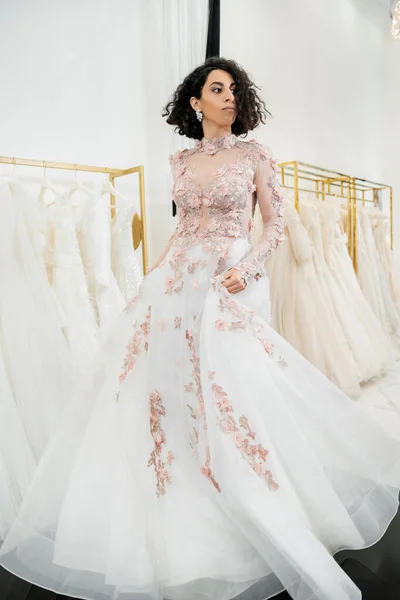 Delightful and middle eastern woman with wavy hair trying on elegant and floral wedding dress inside of luxurious bridal salon, shopping, bride-to-be,  blurred white gown on background — Stock Photo