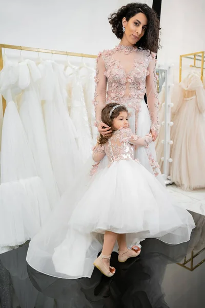 Cute girl in floral attire hugging elegant woman with brunette hair standing in wedding dress near blurred white gown inside of luxurious bridal salon, shopping, bride-to-be, mother and daughter — Stock Photo