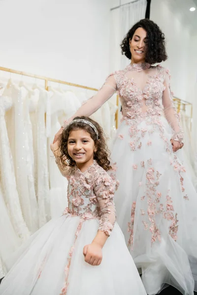 Middle eastern girl in floral attire holding hands with happy woman standing in wedding dress near blurred white gown inside of luxurious bridal salon, shopping, bride-to-be, mother and daughter — Stock Photo