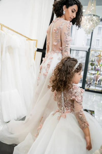 Cute middle eastern girl in floral attire holding hands with woman and walking in floral dresses near blurred wedding gown inside of bridal salon, shopping, bride-to-be, mother and daughter — Stock Photo